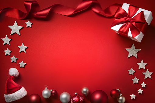 Christmas background in red, with a frame composed of baubles, gift box, stars, ribbon and hat, free illuminated copy space