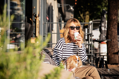 Smiling woman wearing sunglasses and casual clothes while sitting outdoor and drinking morning coffee. Attractive female holding a paper coffee cup in her hand while her cute puppy sitting next to her.