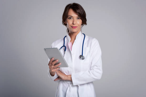 Smiling female doctor with digital tablet standing at isolated grey background stock photo