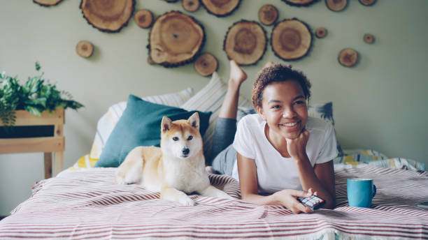 istock Cheerful African American student is watching TV holding remote and pressing buttons choosing television channels while her dog is moving on bed at home. 1436152105