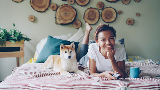 Cheerful African American student is watching TV holding remote and pressing buttons choosing television channels while her adorable dog is moving on bed at home.