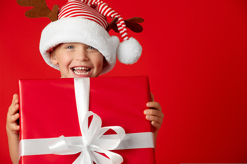 Happy toddler in striped hat with deer cones holds a large red gift box. Child received a gift for Christmas and is happy