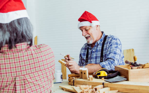 istock Selective focus senior Caucasian retired old man wearing santa hat, check shirt with apron, painting wooden figure as hobby after retirement, celebrating Christmas, New Year, blur foreground friend. 1436155139