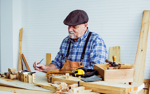 Senior old aging beard kind Caucasian man or carpenter wearing check shirt, apron, making DIY wooden toys for kids, smiling with happiness, sitting alone at indoor home. Retirement, Hobby Concept