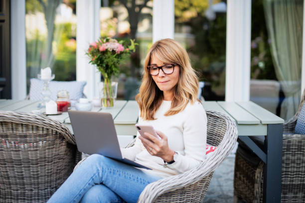 Smiling middle aged woman using text messaging and using laptop on balcony stock photo