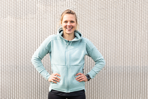 Sportive woman portrait, smiling blonde athlete - happy young woman looking at camera and smiling, gray urban background, wearing pale blue hoodie - sport, success and happiness concepts