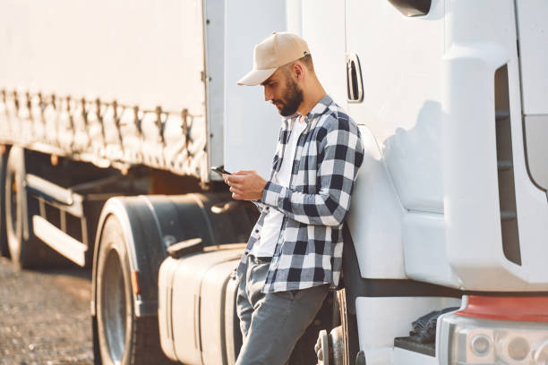Standing and holding smartphone. Young truck driver is with his vehicle at daytime stock photo