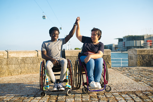 Portrait of supportive biracial family on romantic date. African American man and Caucasian woman in wheelchairs on embankment, raising hands. Love, relationship, happiness concept
