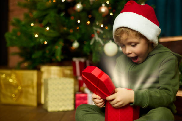 istock Boy in Christmas hat opens a magical gift sitting at a decorated Christmas tree 1436160930