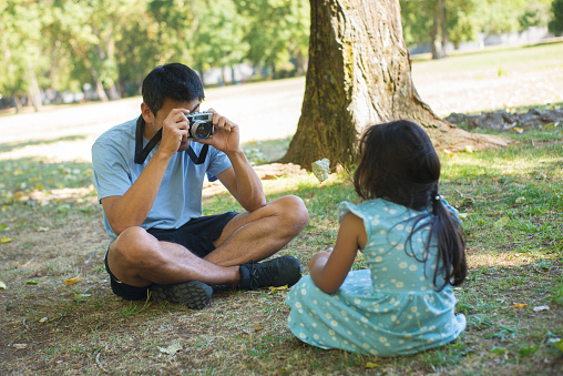 Happy Asian father taking pictures of his beautiful daughter. Young father sitting on grass in city park holding photo camera and little girl looking at camera. Parenthood and togetherness concept