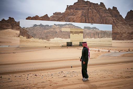 Full length side view of young West Asian man in traditional clothing looking at reflection in mirrored exterior of Maraya Concert Hall in Ashar Valley, Al-Ula.