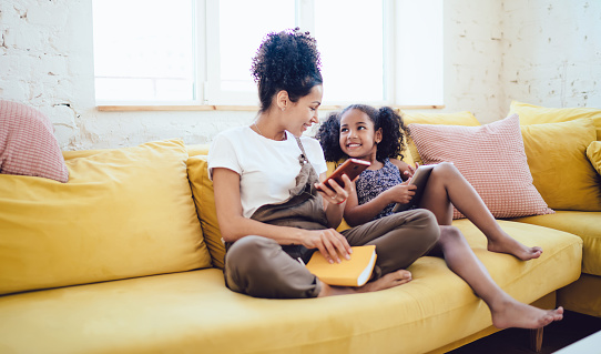 Smiling young black woman holding smartphone and book speaking with happy little daughter with tablet while sitting on yellow sofa in light living room