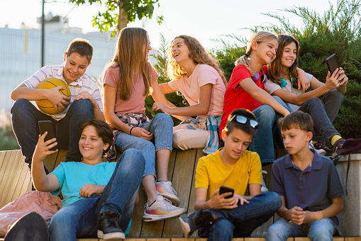 Group of happy teenagers using mobile phones while sitting on the bench in the city