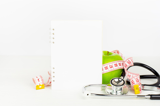 Notepad, green apple with measurement tape and stethoscope on white table. Health eating and dieting concept