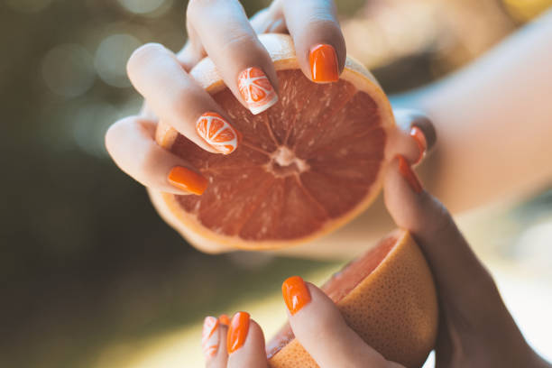 Gentle female hands with a themed manicure hold a grapefruit cut in half. Artistic fingernail painting concept. Sun glare and blurred background, close-up with selective focus