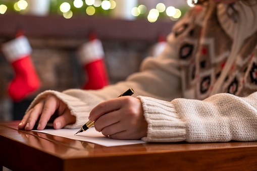 Hand of young woman with fountain pen and sheet of paper on background of christmas fireplace with decoration of light bulbs. Concept of writing wish list.
