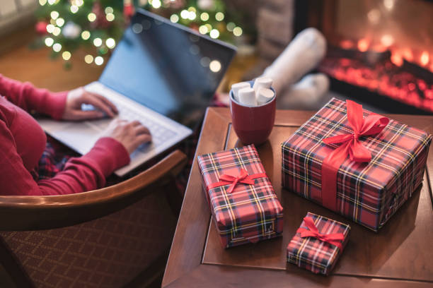 istock Woman holding credit card using laptop for making order sitting at table with packaging gift, cup of cocoa and marshmallows near fireplace and christmas tree. Online shopping concept 1436183384