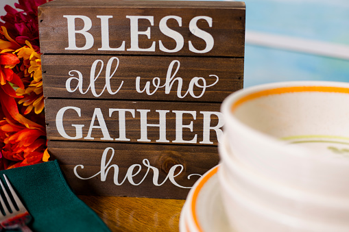 LV Blessing sign on table ready for family gathering during the holidays.  Plates, bowls, napkins and forks are on table in front of orange fall flowers and blessing sign.  Window in background.