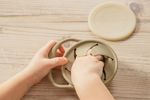 Unrecognizable child taking piece of food out from pastel gray silicone snack cup near lid at wooden table. Baby accessories, tableware, first feeding concept. Top view