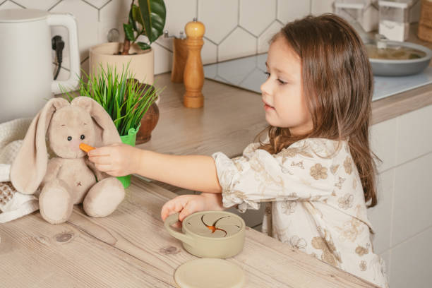 une petite fille aux cheveux noirs de 3-4 ans sort un morceau de carotte d’une tasse à collation en silicone gris pastel et d’un lapin jouet d’alimentation - child caucasian little girls 3 4 years photos et images de collection