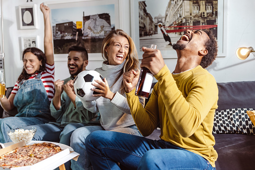 Multiracial group of friends cheering for their sports team and sitting on the sofa in the living room