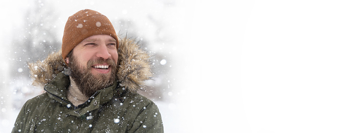 Close up mature man bearded winter coat with fur collar and brown knitted hat snow outdoor. Positive toothy smile looking to the side. Mock up, copy space extra wide banner.