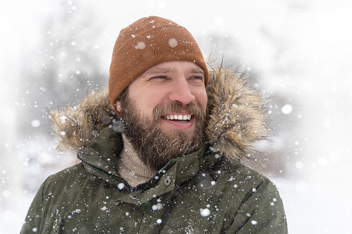Closeup mature man bearded winter coat with fur collar and brown knitted hat snow outdoor. Positive toothy smile looking to the side. Sport and warm clothing concept