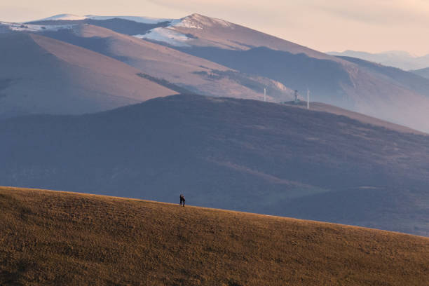 Two distant people on a mountain, with snowcapped peaks and wind turbines.
