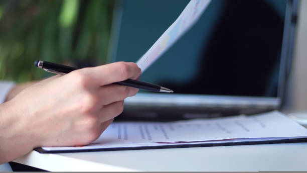 istock Cropped shot of woman's hand writing something while using laptop at desk. close up, on a white desktop there is a white cup of tea, next to a laptop and spread out business graphs 1436216207
