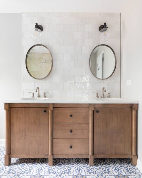 A bathroom with a wood vanity and mosaic tiled floor. A cozy bathroom with a patterned tile floor, natural wood vanity, tiled backsplash, and lights mounted above circular mirrors. egocentric stock pictures, royalty-free photos & images