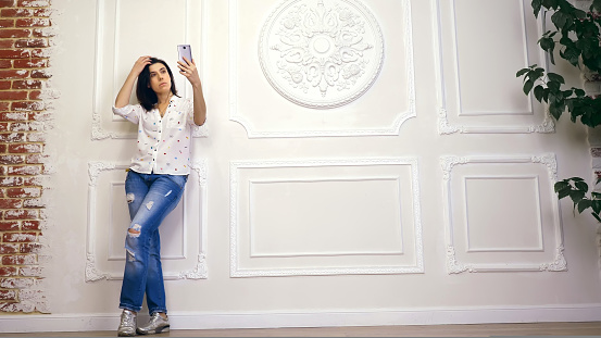 Full length portrait of young woman, girl, brunette, in white shirt and jeans, standing on background of white wall with decorative stucco, with smartphone. looks at screen, takes selfie,. High quality photo