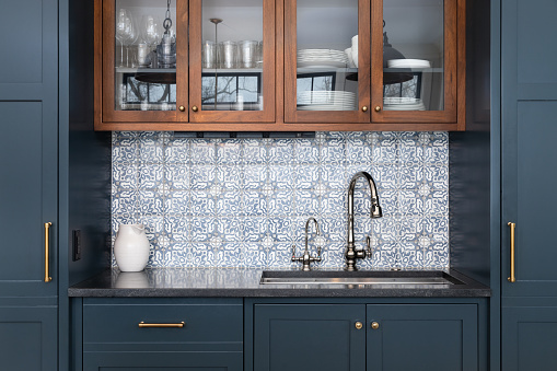 A kitchen sink with a beautiful pattern tiled backsplash with a chrome faucet, black granite countertops, and surrounded by blue and wood cabinets.