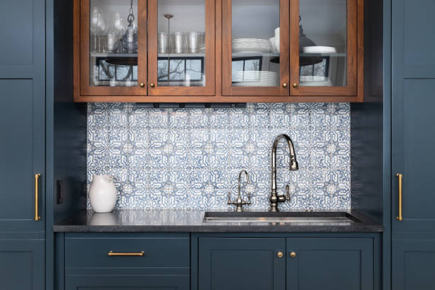 A kitchen with blue cabinets and mosaic tile backsplash. A kitchen sink with a beautiful pattern tiled backsplash with a chrome faucet, black granite countertops, and surrounded by blue and wood cabinets. igneous rock stock pictures, royalty-free photos & images
