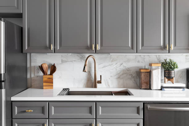 A grey kitchen with a gold faucet and marble subway tile. A kitchen sink detail shot with grey cabinets, a white marble countertop and backsplash, and decorations. cupboard stock pictures, royalty-free photos & images