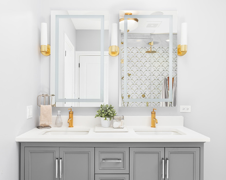 A bright, modern bathroom with a grey vanity cabinet, gold faucets and light fixtures, granite countertop, and a view to a gorgeous tiled walk-in shower.
