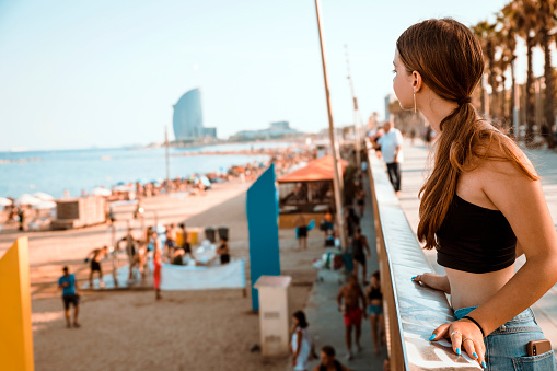 Teenage Girl at Barcelona City waterfront looking at sand beach with people on sunny day. Copy space