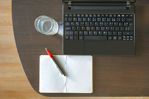 Directly above view of a desk with a laptop, note pad, pen and a glass of water on it.