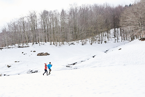 Sportspeople in bright activewear running together near snowy hills with leafless trees during fitness workout on cold winter day in countryside