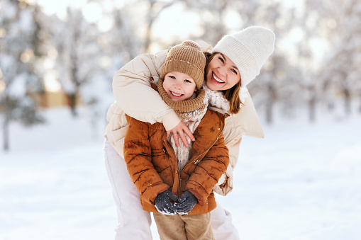 Lovely happy family, mother and little boy son have fun during snowy weather outdoor. Young positive mom embrace kid while playing on fresh frosty air in winter park, selective focus