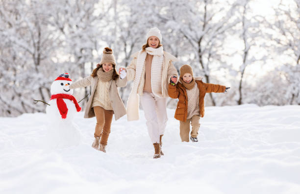 幸せな家族の母親と子供たちは、公園での冬の散歩で雪だるまの近くで遊んだり、走ったり、楽しんだりしています - fun walk ストックフォトと画像