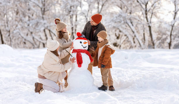 Happy children sculpting funny snowman together with parents in winter snow-covered park Happy parents and children gathering in snow-covered park together sculpting funny snowman from  snow. Father, mother and two kids playing outdoor in winter forest. Family active holiday children in winter stock pictures, royalty-free photos & images