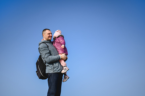 Standing man with a baby girl in arms hugging on a beach with blue sky background. Father hugging daughter.