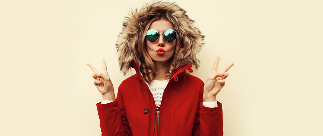 Portrait of beautiful young woman blowing her lips with red lipstick sending sweet air kiss wearing jacket with fur hood on blank background