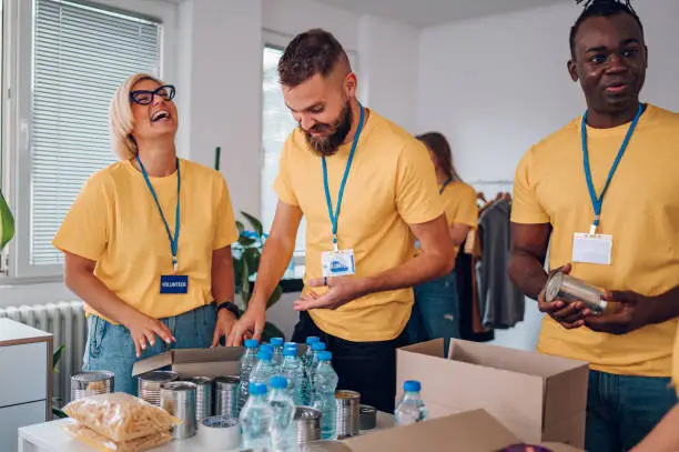 Diverse group of people smiling and working in charitable foundation. Team of multiracial volunteers in yellow uniform packing food in cardboard boxes and working together on donation project indoors.