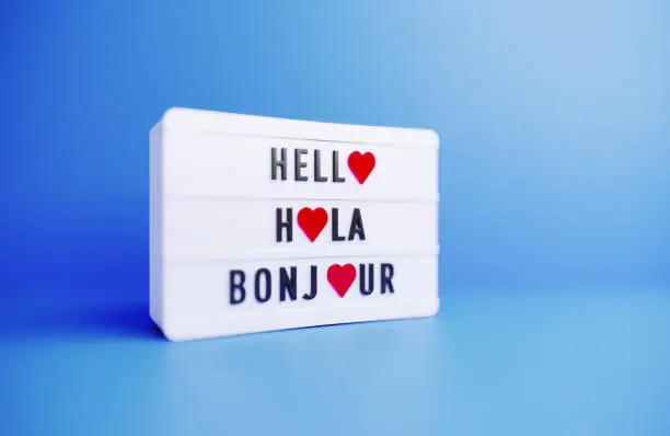 Hello Hola Bonjour written white lightbox sitting on blue background. Horizontal composition with copy space.