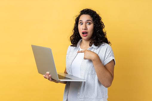 Portrait of amazed astonished woman with dark wavy hair standing with laptop in hands and pointing at display, looking with big eyes and open mouth. Indoor studio shot isolated on yellow background.