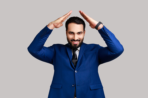 Smiling bearded man standing raising hands showing roof gesture and smiling contentedly, dreaming of house, wearing official style suit. Indoor studio shot isolated on gray background.