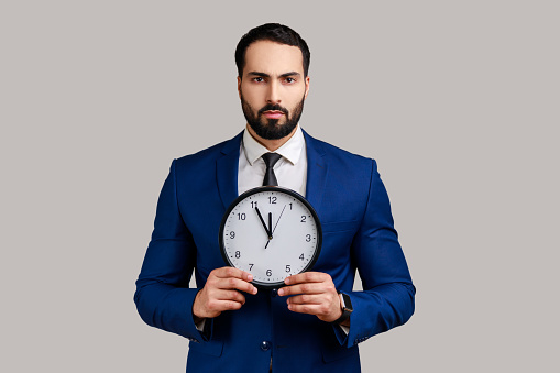 Portrait of serious bossy bearded man afraid of being late, holding in hand wall watch, deadline, punctuality, wearing official style suit. Indoor studio shot isolated on gray background.