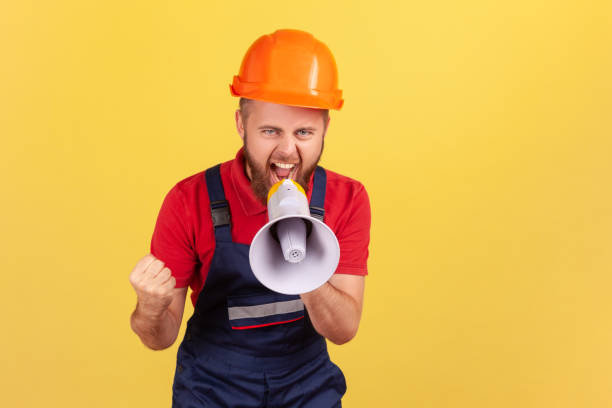 istock Aggressive bearded worker wearing protective helmet and blue overall holding megaphone and screaming 1436258792