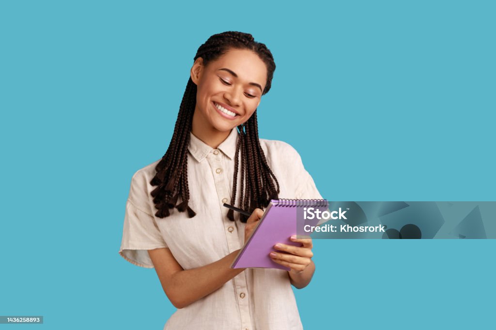 Inspired woman smiling and writing down interesting idea in notebook, making plans, to-do list. Portrait of happy inspired woman with dreadlocks smiling and writing down interesting idea in notebook, making plans, to-do list, wearing white shirt. Indoor studio shot isolated on blue background. People Stock Photo